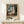 May include: Framed art print showing a woman in white dress standing by a window with blue curtains in an impressionistic style. The poster features text identifying it as "The Girl by the Window" by Edvard Munch from 1893, described as a masterpiece of Expressionism. The wooden-framed print leans against a white wall on a light wooden shelf, with a small wrapped gift box beside it.