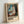 May include: Framed art print leaning against white wall showing Edvard Munch's expressionist painting of a figure in white clothing standing by a window with blue curtains. The artwork features bold brushstrokes in blues, whites, and earth tones. Light wooden frame with cream matting displays the image above black text identifying the piece and artist. Natural lighting creates soft shadows on the wall.