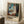 May include: Framed art print leaning against white wall on textured rug, showing Expressionist painting of figure in white dress by window with blue curtains. Warm natural lighting creates shadows on wall. Coffee cup and saucer visible on wooden surface nearby. Light wood frame with descriptive text below artwork.