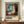 May include: Framed poster print featuring Edvard Munch's expressionist painting of a woman in white nightgown standing by a window with blue curtains. The artwork shows soft, swirling brushstrokes in blues, whites, and earth tones. Large brown-framed print displayed on wooden sideboard alongside white ceramic vase and stacked books against cream wainscoted wall.