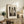 May include: Framed beige and black art print of two embracing figures displayed on rustic wooden sideboard next to fern plant, books, and ceramic vase in neutral-toned living room setting with cream chair and throw blanket