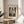 May include: Framed art print displaying Edvard Munch's "The Kiss" in black and white sketch style, showing two embracing figures by a window. Print has cream background with artist name at top and artwork title below. Thin gold or brass frame sits on round wooden side table next to beige upholstered chair. Soft natural lighting creates shadows on neutral wall, with plant leaves visible in corner.