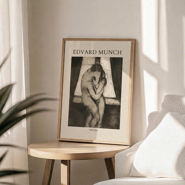 May include: Framed art print displaying Edvard Munch's "The Kiss" in black and white sketch style, showing two embracing figures by a window. Print has cream background with artist name at top and artwork title below. Thin gold or brass frame sits on round wooden side table next to beige upholstered chair. Soft natural lighting creates shadows on neutral wall, with plant leaves visible in corner.