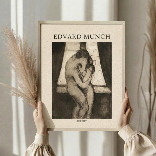 May include: Person in cream outfit holding framed art print in light wooden frame. The artwork shows Edvard Munch's "The Kiss" - a black and white sketch of two figures embracing by a window. Soft natural lighting illuminates neutral-toned room with dried pampas grass visible to the left. The print has cream matting with "EDVARD MUNCH" at top and "THE KISS" at bottom in black text.