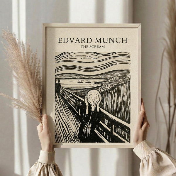 May include: Person in cream blouse holding framed poster print of Edvard Munch's The Scream artwork in light wooden frame. Black line art reproduction on beige background showing iconic screaming figure on bridge with wavy sky. Dried pampas grass visible to left, soft neutral curtains in background create minimalist styled room setting.