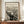 May include: Framed black and white reproduction of Munch's famous expressionist artwork showing a figure with hands to face in an anguished scream, standing on a bridge with swirling sky lines. The print has cream background with dark linear details, displayed in thin brown frame on wooden sideboard alongside white ceramic vase and stacked books in neutral-toned room with white walls and crown molding.
