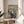 May include: A framed reproduction of Munch's famous expressionist artwork showing a pale figure with hands to face in an anguished scream, standing on a wooden bridge with two silhouettes in background under swirling sky lines. The print sits on a light wood round side table next to a beige upholstered chair in a bright room with white curtains and plant leaves visible.