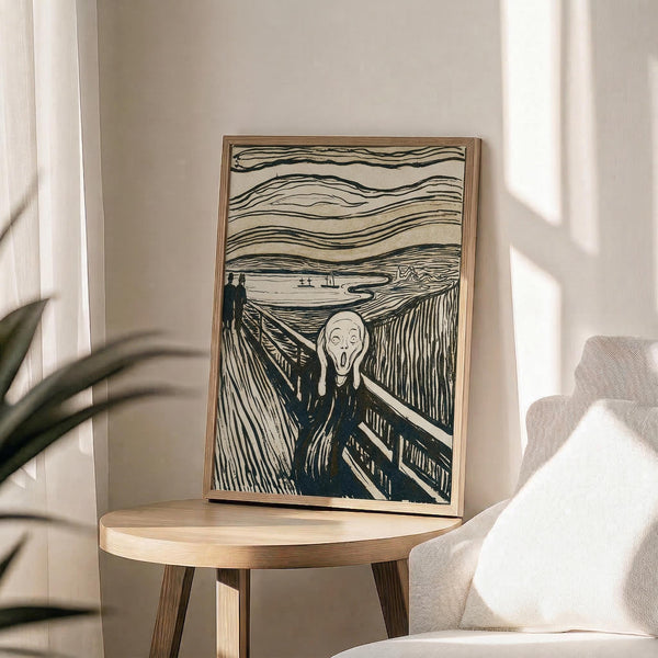 May include: A framed reproduction of Munch's famous expressionist artwork showing a pale figure with hands to face in an anguished scream, standing on a wooden bridge with two silhouettes in background under swirling sky lines. The print sits on a light wood round side table next to a beige upholstered chair in a bright room with white curtains and plant leaves visible.