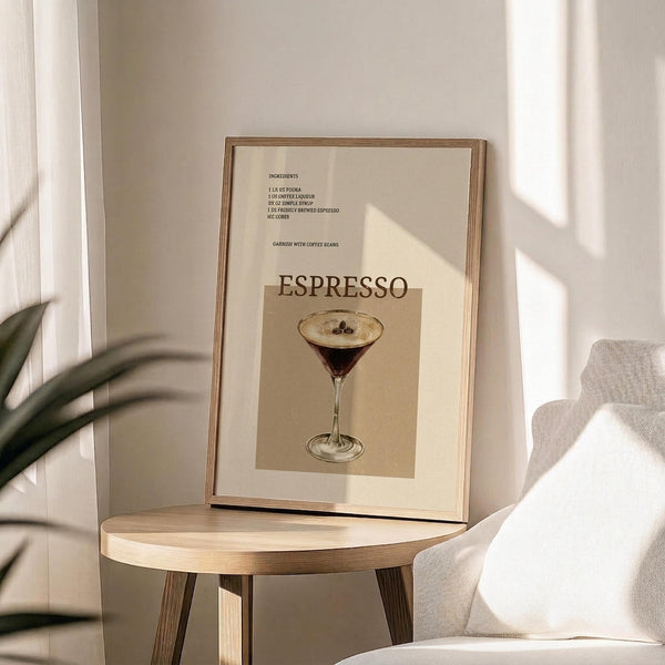 May include: Framed espresso martini recipe poster displayed on round wooden side table next to beige upholstered chair. Print features vintage-style illustration of cocktail in martini glass with coffee beans, cream and brown color palette. Natural lighting creates soft shadows on neutral wall. Green plant leaves visible in corner.