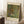 May include: A framed museum-style poster reproduction of Gustav Klimt's colorful garden painting featuring vibrant sunflowers, wildflowers, and lush green foliage in his distinctive decorative style. The cream-colored poster with black text is displayed in a light wooden frame, leaning against white walls on a textured white rug beside a cozy armchair and wooden side table with coffee cup.