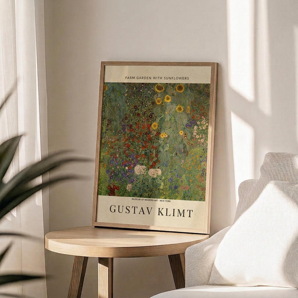 May include: Framed art print displaying a colorful garden scene with sunflowers, wildflowers, and lush greenery in impressionist style, positioned on a round wooden side table next to a beige upholstered chair. Warm natural lighting creates soft shadows on neutral wall. Plant leaves visible in corner of minimalist room setting.