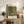 May include: Framed Gustav Klimt reproduction print leaning against white wall on rustic wooden sideboard, showing vibrant garden scene with yellow sunflowers, red poppies, and lush green foliage in impressionist style. Styled with fern plant, ceramic vase, books, and cream upholstered chair with brown leather pillow in warm-toned living room setting.