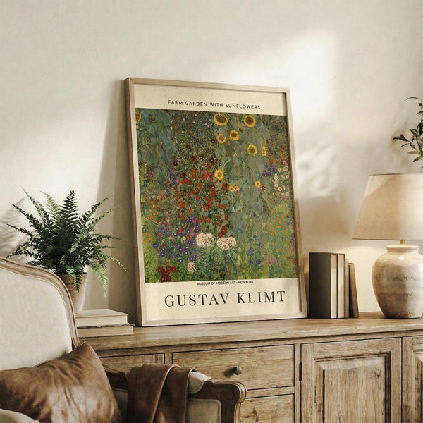 May include: Framed Gustav Klimt reproduction print leaning against white wall on rustic wooden sideboard, showing vibrant garden scene with yellow sunflowers, red poppies, and lush green foliage in impressionist style. Styled with fern plant, ceramic vase, books, and cream upholstered chair with brown leather pillow in warm-toned living room setting.