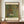 May include: Framed art print displayed on wooden sideboard next to white ceramic vase and stacked books. Print shows colorful garden scene with vibrant sunflowers, red poppies, purple and pink wildflowers in impressionist style. Text reads "Farm Garden with Sunflowers" at top and "Gustav Klimt" at bottom with museum attribution. Warm beige walls and white trim molding visible in background.