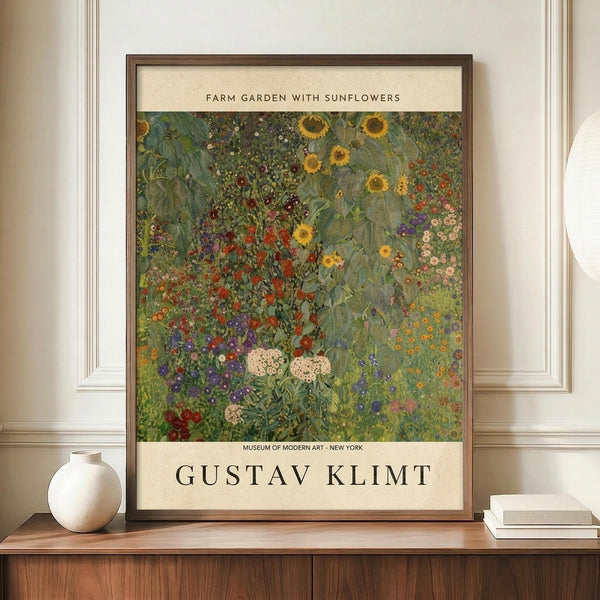 May include: Framed art print displayed on wooden sideboard next to white ceramic vase and stacked books. Print shows colorful garden scene with vibrant sunflowers, red poppies, purple and pink wildflowers in impressionist style. Text reads "Farm Garden with Sunflowers" at top and "Gustav Klimt" at bottom with museum attribution. Warm beige walls and white trim molding visible in background.