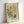 May include: Framed botanical art print with sage green background featuring yellow daisy-like flowers, green foliage, and white leafy branches. Text reads "FLOWER MARKET" at top and "FLORANCE" at bottom with "Papiers Découpés No. 16" in corner. Light wood frame leaning against white wall on light surface with soft natural lighting.