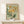 May include: Vintage-style botanical poster in light wood frame featuring yellow daisy-like flowers and green foliage against sage green background with "FLOWER MARKET" and "FRANCE" text, displayed on white wooden surface next to small wrapped gift with twine and dried botanicals against cream wall