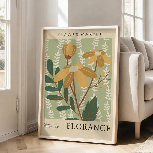 May include: Framed botanical poster with sage green background featuring stylized yellow flowers, white foliage, and dark green leaves in vintage illustration style. Text reads "FLOWER MARKET" and "FLORANCE" with "Papiers Découpés No. 16". Light wood frame leans against white wall next to beige upholstered sofa in bright, naturally lit living room with hardwood floors.