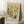 May include: Vintage-style botanical poster with sage green background featuring yellow daisy flowers and dark green foliage, displayed in light wood frame leaning against white wall corner. Scene includes white coffee cup on wooden block and textured cream blanket on white area rug, creating cozy home decor setting with natural window light.