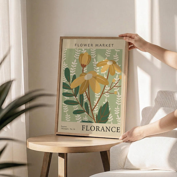 May include: Person's hands holding a framed vintage-style botanical poster featuring yellow daisy-like flowers with green foliage on a sage green background, with "FLOWER MARKET" and "FLORANCE" text. The wooden-framed print sits on a round wooden side table next to a beige upholstered chair, with natural lighting and a potted plant visible in the background.