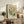 May include: Vintage botanical poster with yellow flowers and green foliage displayed in light wood frame on rustic wooden sideboard. Scene shows cozy living room setting with cream armchair, fern plant, stacked books, ceramic vase, and neutral home decor. Poster features retro flower market design with sage green background and decorative text.