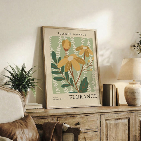 May include: Vintage botanical poster with yellow flowers and green foliage displayed in light wood frame on rustic wooden sideboard. Scene shows cozy living room setting with cream armchair, fern plant, stacked books, ceramic vase, and neutral home decor. Poster features retro flower market design with sage green background and decorative text.