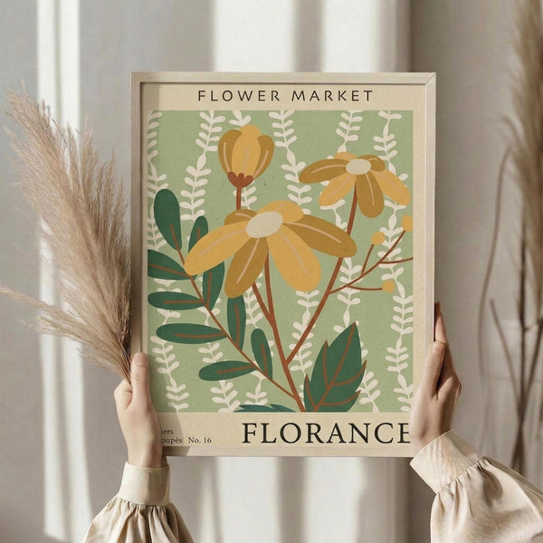 May include: Person in beige ruffled sleeves holding framed botanical poster featuring yellow daisy-like flowers and green foliage on sage green background with "Flower Market" and "Florance" text, alongside dried pampas grass stems, against soft neutral curtained window setting