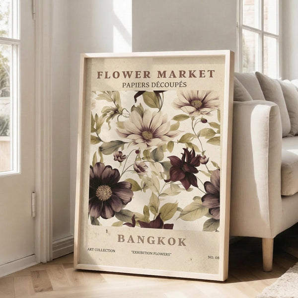 May include: Large framed botanical poster leaning against white wall next to beige armchair. Vintage-style print features cream and burgundy flowers with green leaves on aged beige background. Text reads "Flower Market," "Papiers Decoupes," and "Bangkok" with "Art Collection Exhibition Flowers No. 08." Light wood frame, bright room with windows and hardwood floors.