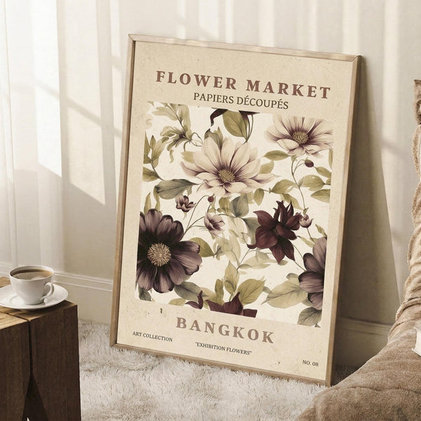 May include: A framed botanical poster displaying vintage-style illustrations of purple and white flowers with green foliage on cream background, text reading "Flower Market" and "Bangkok" at top and bottom. The wooden-framed print leans against white wainscoting beside a wooden side table with white coffee cup and saucer, soft beige throw blanket visible, natural window light creating shadows.