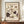 May include: Vintage-style botanical poster with cream background featuring purple and white flowers with green leaves, displayed in brown wooden frame on wood sideboard next to white ceramic vase and stacked books against white paneled wall