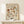 May include: Framed botanical poster with vintage-style floral illustration in warm earth tones including rust, amber, and sage green flowers and leaves on cream background, displayed on light wood console table with small white wrapped gift box beside it against neutral wall