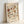 May include: Framed botanical poster with vintage-style floral illustrations in warm earth tones including rust, orange, and sage green flowers and leaves against cream background, displayed in light wood frame leaning against white wall with soft natural lighting