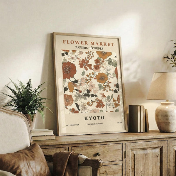 May include: Framed botanical poster with vintage-style floral illustrations in warm earth tones including rust, orange, and sage green flowers and leaves on cream background. Print displays on rustic wooden sideboard alongside fern plant, books, ceramic vase, and table lamp in cozy neutral-toned living room setting with leather furniture.