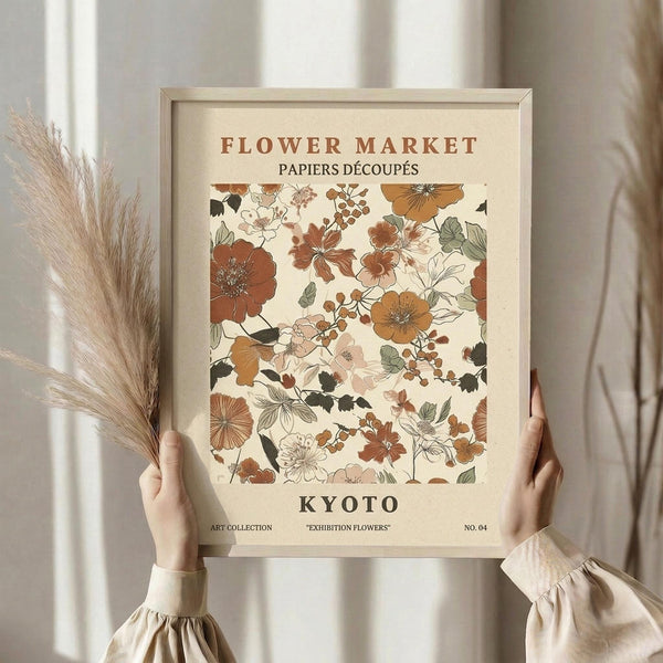 May include: Person in cream ruffled blouse holding framed botanical poster featuring vintage-style flowers in rust, orange, and sage green tones. Light wood frame against cream textured background with dried pampas grass stems visible on left side. Poster shows floral pattern with "FLOWER MARKET" and "KYOTO" text in earth tone color palette.