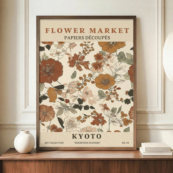 May include: Framed vintage-style botanical poster displaying various flowers in warm earth tones including rust, sage green, and cream colors. The artwork features "FLOWER MARKET" and "PAPIERS DÉCOUPÉS" at top, "KYOTO" prominently in center, with "ART COLLECTION" and "EXHIBITION FLOWERS" at bottom. Dark wooden frame sits on light wood sideboard next to white ceramic vase and stack of books against cream wall with white trim molding.
