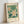 May include: Framed botanical poster print leaning against white wall, featuring stylized white flowers with yellow centers on green leafy background, displayed in light wood frame on cream mat with "FLOWER MARKET" and "MADRID" text, soft natural lighting creating gentle shadows