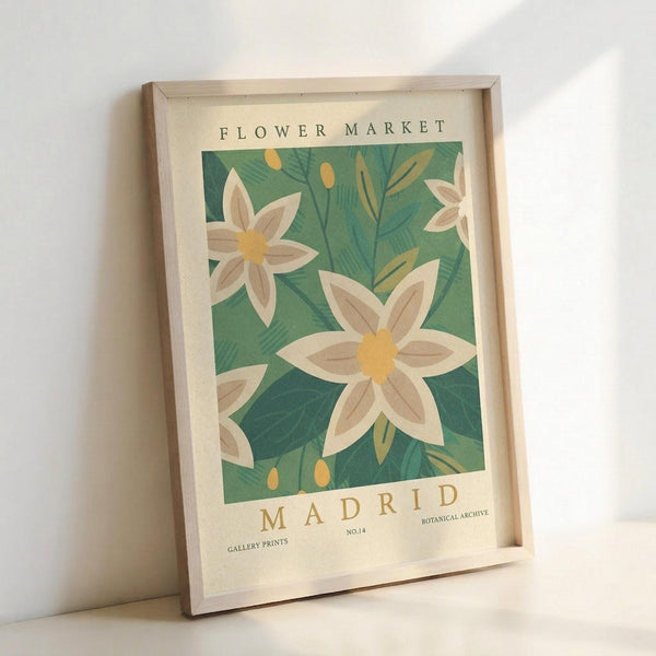 May include: Framed botanical poster print leaning against white wall, featuring stylized white flowers with yellow centers on green leafy background, displayed in light wood frame on cream mat with "FLOWER MARKET" and "MADRID" text, soft natural lighting creating gentle shadows