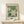 May include: Framed botanical poster with white flowers and yellow centers on teal green background, displayed on light wood console table against cream wall. Small wrapped gift with evergreen sprig sits nearby. Natural wood frame with glass, vintage-inspired floral design in muted earth tones.