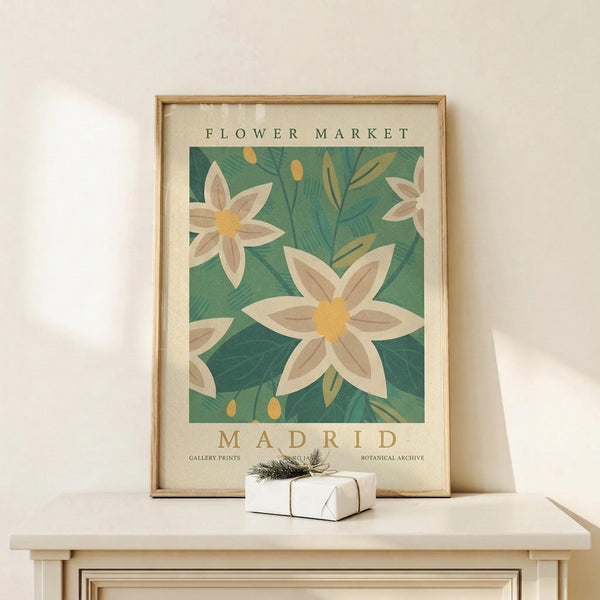 May include: Framed botanical poster with white flowers and yellow centers on teal green background, displayed on light wood console table against cream wall. Small wrapped gift with evergreen sprig sits nearby. Natural wood frame with glass, vintage-inspired floral design in muted earth tones.