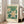 May include: Framed botanical poster with white flowers and yellow centers against green leafy background, displayed in bright living room corner next to beige upholstered chair. Natural wood frame rests on light hardwood floor near tall white-trimmed windows with soft sunlight streaming in.