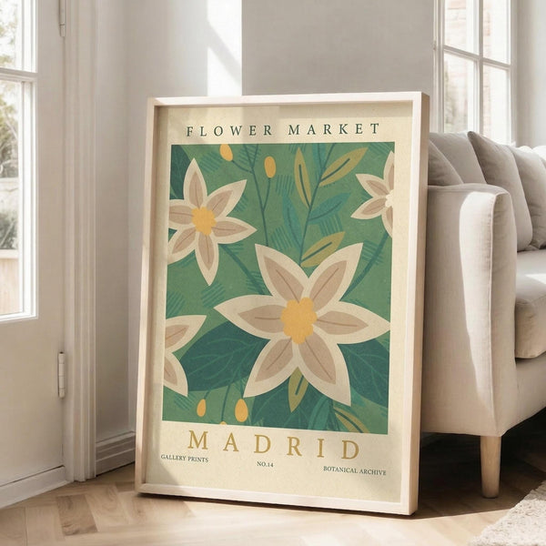 May include: Framed botanical poster with white flowers and yellow centers against green leafy background, displayed in bright living room corner next to beige upholstered chair. Natural wood frame rests on light hardwood floor near tall white-trimmed windows with soft sunlight streaming in.
