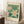 May include: Vintage-style botanical poster with white flowers and yellow centers on green leafy background, displayed in light wood frame leaning against white wall corner. Scene shows cozy interior with white coffee cup on wooden block, cream textured rug, and beige throw blanket. Natural window light creates soft shadows on wall.