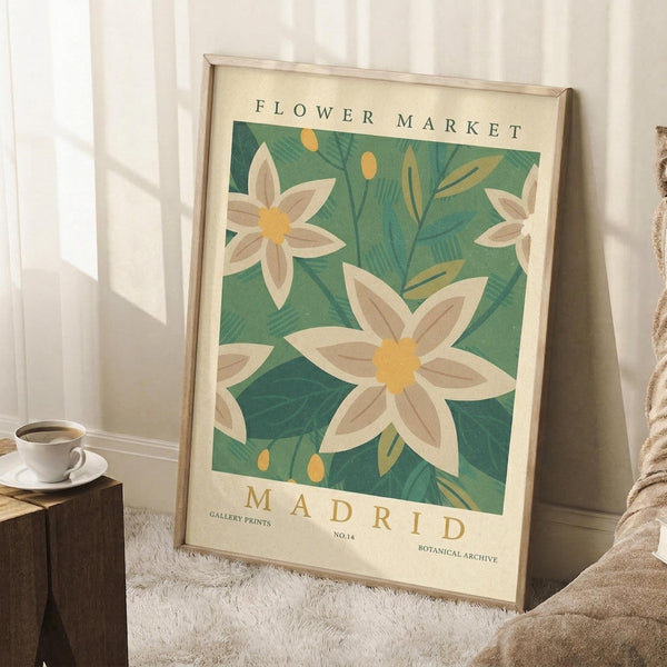 May include: Vintage-style botanical poster with white flowers and yellow centers on green leafy background, displayed in light wood frame leaning against white wall corner. Scene shows cozy interior with white coffee cup on wooden block, cream textured rug, and beige throw blanket. Natural window light creates soft shadows on wall.
