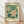 May include: Framed botanical art print leaning against white wall on light wood floor. Features cream-colored poster with green leafy background and white flowers with yellow centers. Text reads "Flower Market" at top and "Madrid" at bottom. Natural wood frame with warm lighting and soft shadows across the scene.