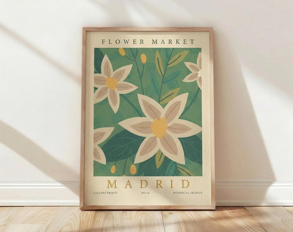 May include: Framed botanical art print leaning against white wall on light wood floor. Features cream-colored poster with green leafy background and white flowers with yellow centers. Text reads "Flower Market" at top and "Madrid" at bottom. Natural wood frame with warm lighting and soft shadows across the scene.