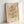 May include: Framed botanical poster print in light wood frame leaning against white wall. Artwork features stylized flowers with orange and yellow blooms, green stems, scattered leaves and small botanical elements on peachy beige background. Text reads "FLOWER MARKET" at top and "NEW YORK GALLERY PRINTS MUSEUM OF MODERN ART NO.5" at bottom in brown lettering. Warm, earthy color palette with mid-century modern illustration style.
