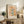 May include: Framed vintage-style botanical poster with orange and yellow flowers on peachy background, displayed on rustic wood sideboard alongside fern plant, stacked books, ceramic vase and table lamp in warm neutral living room setting