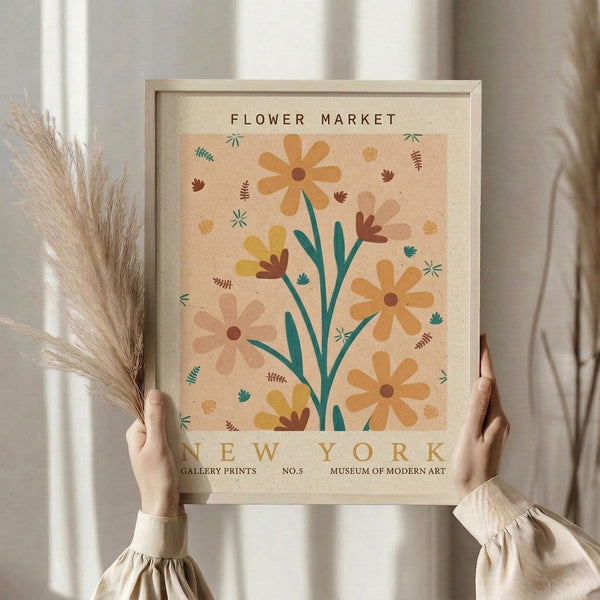 May include: Person in cream ruffled sleeves holding framed botanical poster with stylized flowers in warm oranges, yellows, and teals on beige background. Light wood frame against white curtained window. Dried pampas grass held in other hand. Soft natural lighting creates cozy, minimalist aesthetic.