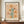 May include: Framed vintage-style botanical poster with peachy beige background featuring stylized flowers in orange, yellow, and brown tones with teal green stems and leaves. Small decorative botanical elements scattered throughout. Text reads "FLOWER MARKET" at top and "NEW YORK" at bottom with museum gallery details. Dark wooden frame displayed on wooden sideboard next to white ceramic vase and stacked books in bright, modern room with white paneled walls.