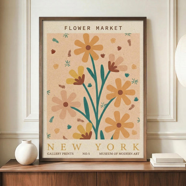 May include: Framed vintage-style botanical poster with peachy beige background featuring stylized flowers in orange, yellow, and brown tones with teal green stems and leaves. Small decorative botanical elements scattered throughout. Text reads "FLOWER MARKET" at top and "NEW YORK" at bottom with museum gallery details. Dark wooden frame displayed on wooden sideboard next to white ceramic vase and stacked books in bright, modern room with white paneled walls.